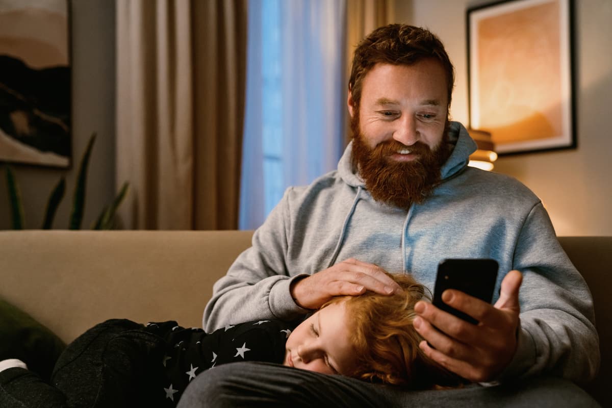 Father with beard looking at phone while his child rests on his lap